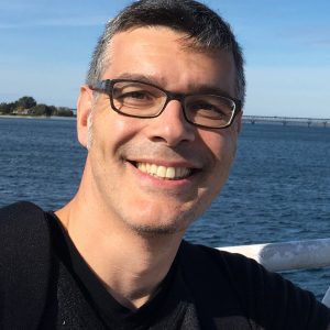 Man with short dark gray hair and black glasses on a bridge over a body of water